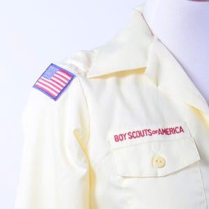 以下が含まれることがあります： 左肩に青と赤のアメリカ国旗のパッチと、赤い刺繍で「BOY SCOUTS of AMERICA」と書かれたパッチが付いた、白い長袖シャツです。