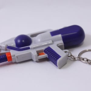 May include: A miniature water gun keychain in shades of gray, purple, and orange. The toy gun features a trigger, nozzle, and a ring for attaching keys. The water gun is a novelty item.