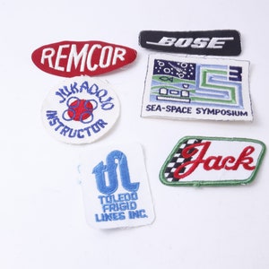 May include: A collection of seven embroidered patches, including a red and white "REMCOR" patch, a white patch with blue text that says "JURADO INSTRUCTOR", a black and white "BOSE" patch, a white patch with blue and green text that says "SEA-SPACE SYMPOSIUM", a green and white patch with red text that says "Jack", a white patch with blue text that says "TOLEDO FRIGID LINES INC.", and a white patch with red text that says "JURADO INSTRUCTOR".