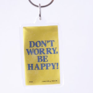 Puede incluir: Un llavero rectangular con fondo amarillo y el texto azul "DON'T WORRY, BE HAPPY!". El llavero es de plástico transparente y tiene un aro de metal plateado en la parte superior.