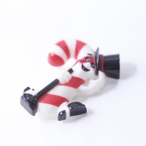 Könnte beinhalten: Eine weiße und rote Zuckerstangenfigur, die einen schwarzen Zylinder trägt. Die Figur hält einen schwarzen Stock und hat ein lächelndes Gesicht.