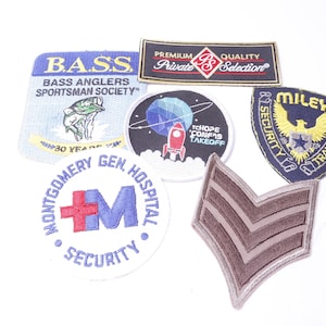 Może przedstawiać: Zbiór haftowanych naszywek, w tym niebieska i biała naszywka z tekstem "BASS ANGLERS SPORTSMAN SOCIETY", niebieska i biała naszywka z rakietą i tekstem "TCHOPE CONFIS TAKEOFF", niebieska naszywka z żółtym orłem i tekstem "MILEY SECURITY TRANSPORT", biała naszywka z czerwonym krzyżem i tekstem "MONTGOMERY GEN HOSPITAL SECURITY", brązowa naszywka z dwoma paskami i czerwono-biała naszywka z tekstem "PREMIUM QUALITY Private Selection".