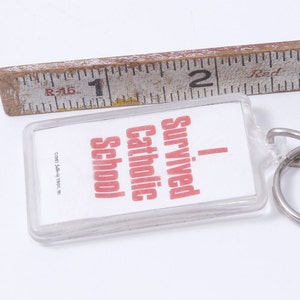 May include: A clear plastic keychain with a white background and red text that reads "I Survived Catholic School".