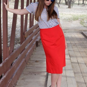 May include: A woman wearing a blue and white patterned blouse with a red skirt and black heels. She is standing on a wooden bridge.