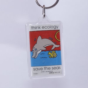 Puede incluir: Un llavero de plástico transparente con una imagen colorida de un delfín saltando fuera del agua con un pez pequeño. El texto en el llavero dice "think ecology save the seas ©1991 Jim Benton LittleHip Troy, MI K1434".