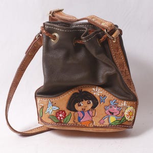 May include: Brown leather bucket bag with a hand-painted design featuring a cartoon girl, a monkey, flowers, and butterflies. The bag has a drawstring closure, a shoulder strap, and decorative stitching.
