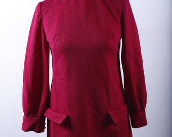 Puede incluir: Un vestido rojo oscuro de manga larga y cuello alto. El vestido tiene una tela texturizada y dos bolsillos grandes en la parte delantera.