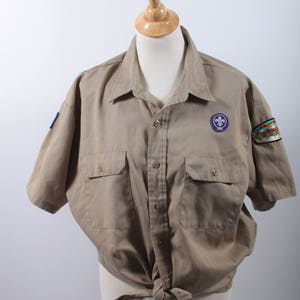 Può includere: Una camicia beige a maniche corte con bottoni, due tasche e un emblema scout sul lato destro. La camicia è annodata in vita.