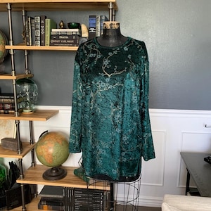 May include: A long-sleeved, emerald green velvet top with a floral pattern in gold and light blue. The top is displayed on a wire dress form. The background includes a bookshelf, globe, and framed artwork.