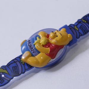 May include: A blue plastic Winnie the Pooh watch with a picture of Winnie the Pooh holding a pot of honey. The watch has the word "Hunny" written on the pot of honey.