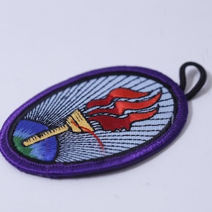 May include: Oval-shaped embroidered patch with a purple border. The patch features a torch with red flames, a blue and green globe, and a light blue background with radiating lines. A black loop is attached.