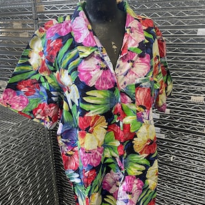 Puede incluir: Una camisa de manga corta con botones y estampado floral colorido. La camisa tiene un fondo negro con flores rosas, rojas, amarillas y azules.