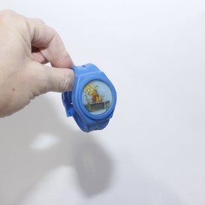 Pu&ograve; includere: Un orologio da polso in plastica blu con display digitale e un'immagine di cartone animato di Bart Simpson sul quadrante.