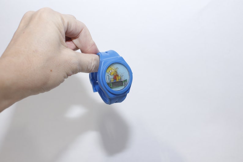 Pu&ograve; includere: Un orologio da polso in plastica blu con display digitale e un'immagine di cartone animato di Bart Simpson sul quadrante.