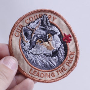 Puede incluir: Un parche bordado beige con una cabeza de lobo en el centro. El texto alrededor del lobo dice "Cub Country Leading the Pack" y una flor de lis roja está en el lado derecho del parche.