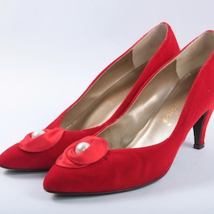 Op de afbeelding: Een paar rode suède pumps met een spitse neus en een hoge hak. Elke schoen heeft een satijnen accent met een parelversiering. De binnenkant van de schoen is goudkleurig.