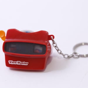 Può includere: Un portachiavi View-Master rosso con una catena e un portachiavi argentati. Il giocattolo classico ha uno schermo di visualizzazione nero e una leva arancione. Le parole "View-Master" sono stampate in bianco sul davanti.