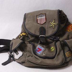 以下が含まれることがあります： オリーブグリーンのキャンバスバックパック。星や盾など、さまざまな刺繍パッチが付いています。黒革のアクセント、フラップ開閉、調節可能なストラップが付いています。複数のポケットと巾着開閉が特徴です。