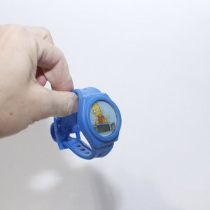 Pu&ograve; includere: Un orologio da polso in plastica blu con un'immagine di cartone animato di Bart Simpson sul quadrante.
