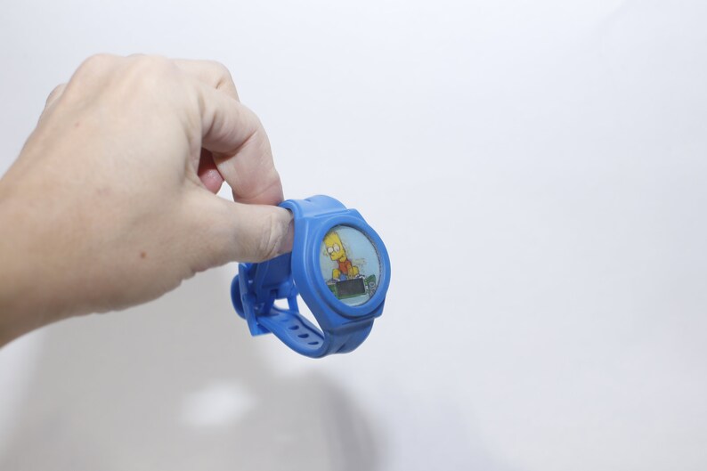 Pu&ograve; includere: Un orologio da polso in plastica blu con un'immagine di cartone animato di Bart Simpson sul quadrante.
