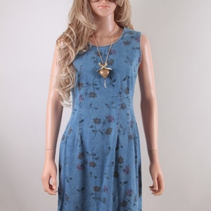 May include: A sleeveless denim dress with a floral print in shades of blue, purple, and green. The dress has a scoop neck and a fitted bodice. A gold heart-shaped pendant hangs from a gold chain necklace.