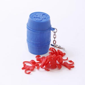 Può includere: Un portachiavi blu a forma di botte in plastica con catena e anello argentati. Diversi piccole scimmie di plastica rossa sono sparse intorno alla botte. La botte ha un coperchio con un disegno.