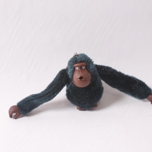 May include: A plush gorilla figurine with outstretched arms. The gorilla has a dark teal body and arms, a brown face, and brown hands and feet. The figurine has a small loop at the top for hanging.