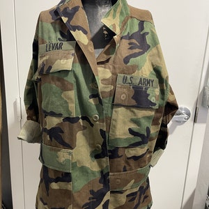 Op de afbeelding: Een groene, bruine en zwarte camouflagejas met knoopsluiting. De jas heeft twee grote zakken aan de voorkant en een kleinere zak op de linkermouw. De woorden "U.S. ARMY" en "LEVAR" staan op de jas gedrukt.
