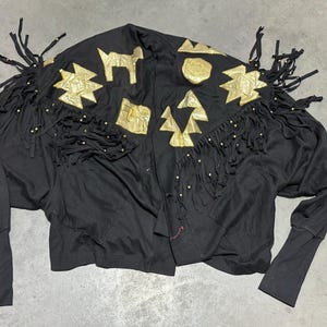 May include: Black jacket with gold-colored geometric shapes and fringe detailing. The jacket features long sleeves and a unique design with gold embellishments and small gold beads. A vintage-inspired piece.