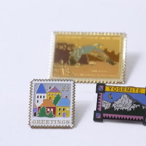 May include: Three enamel pins featuring a colorful house scene, a bird in flight, and a mountain range with the word "Yosemite" in a yellow banner. The pins are all in a silver frame with a white background.