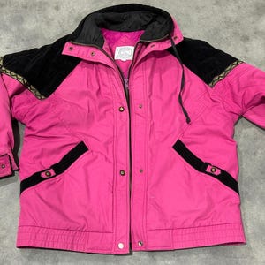May include: A bright pink winter jacket with black accents. The jacket features a black collar, shoulder patches, and pocket details. Gold trim accents the shoulder patches. The jacket has a zipper closure and snap buttons.