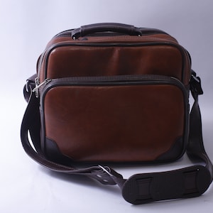 Puede incluir: Un bolso de hombro de cuero marrón con asa superior y correa ajustable. El bolso tiene dos compartimentos con cremalleras.