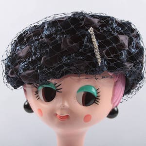 May include: A black and blue satin hat with a net veil and a rhinestone pin. The hat is worn on a doll's head with pink hair and large blue eyes.