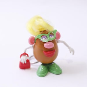 May include: A whimsical Mr. Potato Head toy with a brown body, yellow hair, and green glasses. It has pink ears, a pink nose, and a red mouth. The toy is holding a red purse and wearing green shoes.