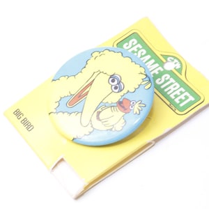 Op de afbeelding: Een gele Sesame Street Big Bird-knop met een blauwe achtergrond. Big Bird houdt een klein geel vogeltje vast met een rode snavel. De knop is bevestigd aan een gele kaart met de tekst "Sesame Street" erop.