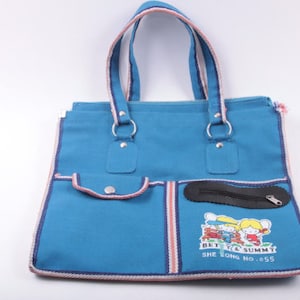 May include: A blue canvas tote bag with white and pink trim. The bag has a zippered pocket and a smaller pocket on the front. It features a cartoon image of two children and the text "Betty & Sammy She Long No. 055".