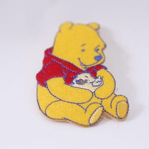Op de afbeelding: Een gele Winnie de Poeh-patch met een rood shirt en blauwe stiksels. De patch toont de beer die een honingpot vasthoudt. De patch is gemaakt van vilt en is geschikt voor kleding of accessoires.