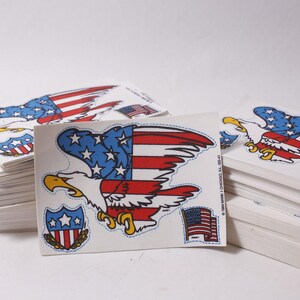 May include: Piles of stickers depicting an American bald eagle with a red, white, and blue flag design. The stickers also include a shield and a small American flag. The stickers are on a white background.