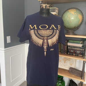 Peut inclure: Un t-shirt bleu marine avec un motif doré d'un aigle stylisé avec le mot "MOAB" au-dessus et "Utah" en dessous.