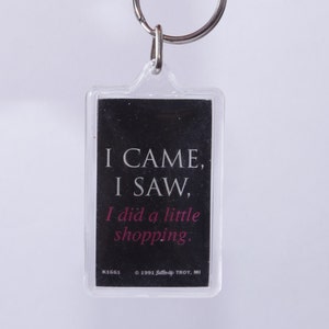 Könnte beinhalten: Ein durchsichtiger Schlüsselanhänger aus Kunststoff mit schwarzem Hintergrund und weißem Text, der "I CAME, I SAW, I did a little shopping." lautet. Der Text "KIGGI © 1991 TROY, MI" ist unten in Weiß gedruckt.