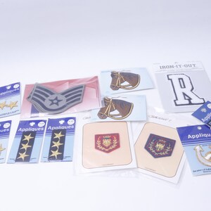 May include: Assortment of iron-on patches, including a military rank patch, a horse head, a letter R, a crest, an anchor, and stars. The patches are packaged on white cards with the word "Appliques" printed on them.
