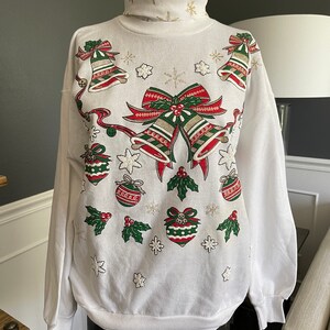 May include: White Christmas sweater with a festive design featuring red and green bells, bows, and ornaments. The sweater has a turtleneck and long sleeves.