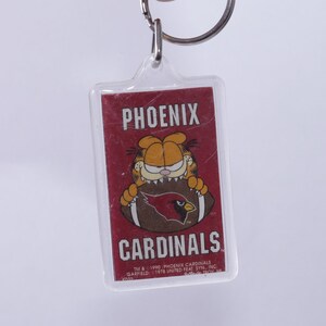 May include: A clear plastic keychain with a red and white image of Garfield the cat holding a football with the Phoenix Cardinals logo. The text "PHOENIX CARDINALS" is printed on the keychain.