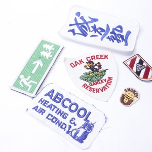 May include: A collection of seven embroidered patches. The patches feature various designs, including a green exit sign, a white patch with the text "ABCooL Heating & Air Cond.", a white patch with the text "Oak Creek Oakley Reservation", a white patch with blue and red text, a white patch with red and black stripes, a brown patch with a bear wearing a ranger hat, and a white patch with a blue and red design.