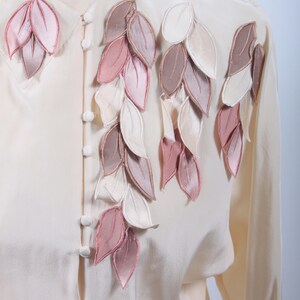 Può includere: Una blusa color crema con un designo decorativo di foglie rosa, marroni e bianche.