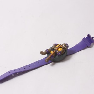 May include: A purple silicone watch strap featuring a small, three-dimensional figure of a Teenage Mutant Ninja Turtle character wearing a yellow chest plate and purple mask, holding a brown staff, attached to a circular black watch face housing.