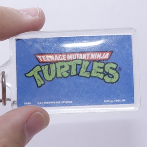 May include: A clear plastic keychain with a blue background and the Teenage Mutant Ninja Turtles logo in green. The text "Teenage Mutant Ninja Turtles" and "K&S®" are printed in white. The text "©1989 Mirage Studios" and "Suttle-Up TROY, MI" are printed in black.
