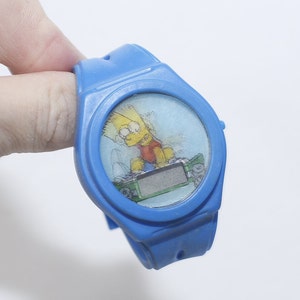 Pu&ograve; includere: Orologio da polso in plastica blu con display digitale e un'immagine di cartone animato di Bart Simpson che fa skateboard.