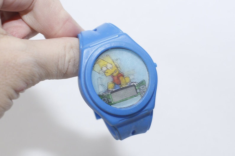 Pu&ograve; includere: Orologio da polso in plastica blu con display digitale e un'immagine di cartone animato di Bart Simpson che fa skateboard.