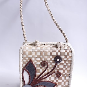 May include: A woven straw handbag with a white braided strap. The bag has a floral design with brown, white, and black accents. The flower has a pearl-like center.